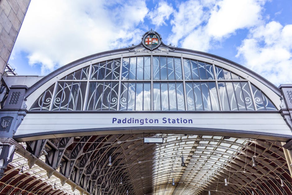 The image shows the ornate archway of Paddington Station with intricate metalwork and glass panels, reflecting the cloudy sky above. Below the arch, there is a white sign with blue text reading 'Paddington Station'. The station's architectural details include decorative elements and a crest at the top of the arch. In the foreground, part of a loading area is visible, with a few cardboard boxes and wrapped furniture suggesting preparations for a home relocation. Sunlight filters through the glass roof structure, illuminating the area where removal and transport activities occur. A removal van may be nearby, associated with Removal Van Paddington, supporting furniture transport and packing services for house moves, particularly in urban narrow-street environments like Norfolk Square. The scene captures a typical logistical setting for moving services, highlighting the station's historic architecture and the ongoing packing and loading process.