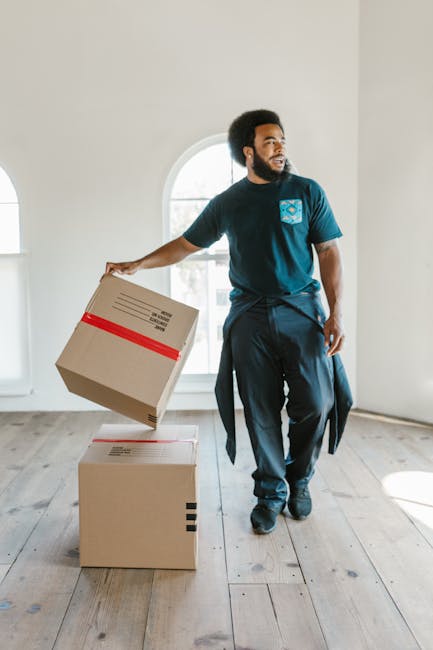 A man with a beard and curly hair, dressed in a navy T-shirt and dark trousers, is standing inside a bright, minimally furnished room with wooden flooring and large arched windows in the background. He is holding a cardboard box with red tape, preparing for a home relocation or furniture transport process, while another box is placed on the floor nearby. The scene captures the packing and moving phase typical of house removals, with natural light illuminating the space. This visual aligns with services provided by Removal Van Paddington for detailed house removals and moving logistics in narrow-street environments, such as the Norfolk Square area referenced in the page title.